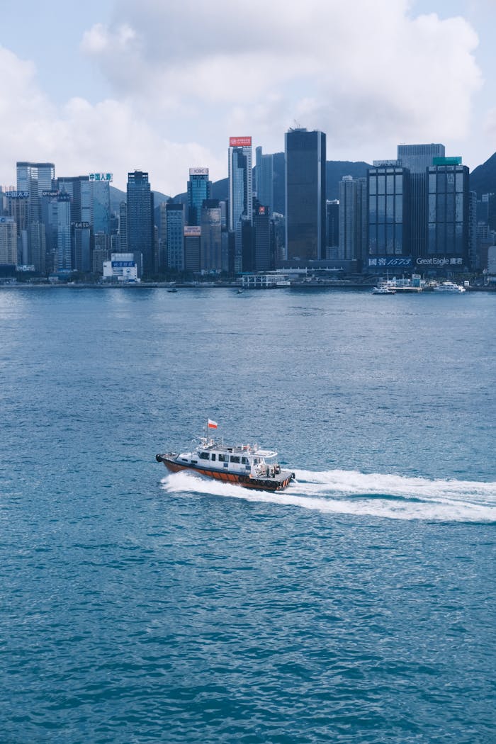 A boat speeds across Hong Kong Harbor with the city skyline in the background. Bright, vibrant urban scene.